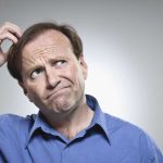 A man in a blue shirt with a confused expression, scratching his head