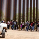 People walking beside tall fence and border patrol vehicle