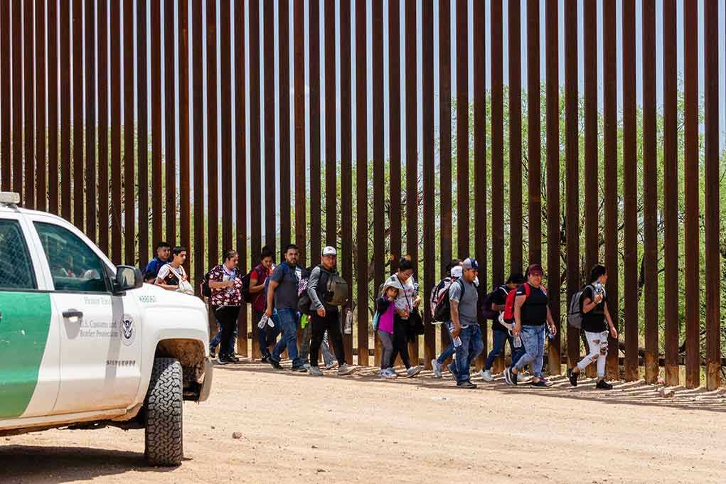 People walking beside tall fence and border patrol vehicle