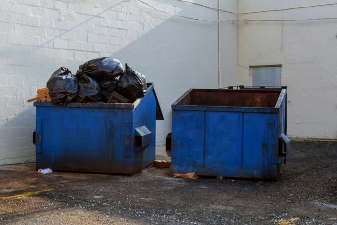 779940112 Two blue dumpsters, one overflowing with trash bags.