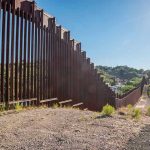 Trump BYPASSES 25 Laws — Wall Rising! Tall metal border wall with rural landscape.