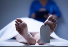 A deceased body on a table with a tag on the foot, and a medical professional in the background