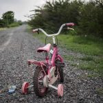 Parents Vanish! — Child Left to FEND FOR HIMSELF Pink childrens bike with training wheels on gravel path.