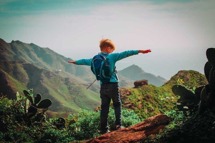 Child with backpack stands on mountain, arms outstretched.