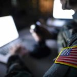 Missile Defense SHOCK Upgrade Reshapes U.S. Power Soldier using laptop with US flag patch visible.