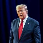 Trump’s Insurrection Act Threat Very Real Man in suit with red tie speaking on stage.