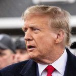 King Charles’s US Visit Sparks Backlash Man in suit and red tie speaking outside.