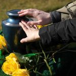 Canada Kills 26-Year-Old for Blindness Hands touching an urn surrounded by yellow roses.