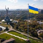 Zelenskiy Bans Russian Church—Religious Freedom Eroded? Large Ukraine flag with cityscape in background.