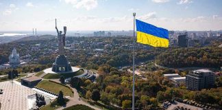 Zelenskiy Bans Russian Church—Religious Freedom Eroded? Large Ukraine flag with cityscape in background.