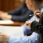 Polite Words Hid DECADES of HORROR Person holding pen, speaking to seated individuals with papers.