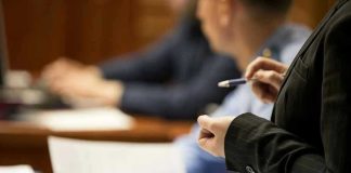 Polite Words Hid DECADES of HORROR Person holding pen, speaking to seated individuals with papers.