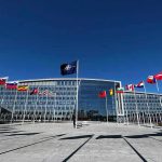 Nuclear Line ERASED — Finland Blinks Flags outside NATO headquarters building under clear blue sky.
