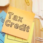HURRY — This TAX-CREDIT is STILL AVAILABLE — Democrats WANT To SQUASH It. Sticky note labeled Tax Credit on a desk with papers and charts