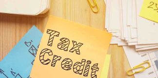HURRY — This TAX-CREDIT is STILL AVAILABLE — Democrats WANT To SQUASH It. Sticky note labeled Tax Credit on a desk with papers and charts
