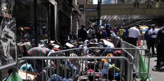 HOMELESS Bureau GUTTED — Where’d $300M Go? A crowded street with individuals sitting behind barriers and bags, highlighting urban homelessness