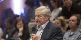 BOMBSHELL — Soros’ BLITZ Warps 2026 Map An elderly man speaking into a microphone at a conference with an attentive audience