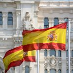 Trump TARGETS Spain — Huge Reason WHY! Two Spanish flags waving in front of a historic building