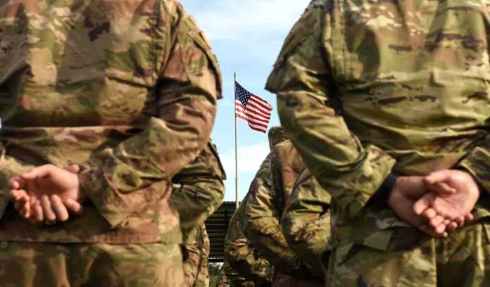 shutterstock_1507031819 Soldiers in camouflage stand with hands behind backs facing a U.S. flag