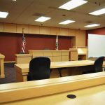 He Shot Three Students On A Sidewalk — Then He Opened His DOOR Interior view of an empty courtroom with wooden furniture and American flags