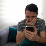 Dad Called And Called — Someone Made Sure NOBODY Came Man sitting on couch looking worried at smartphone