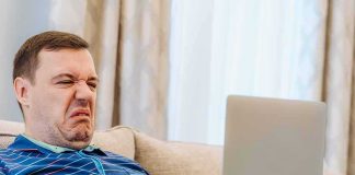 Men’s Blue Pill RECALLED — The THIRD Dangerous Drug HIDDEN Inside Man grimacing while using a laptop on a couch