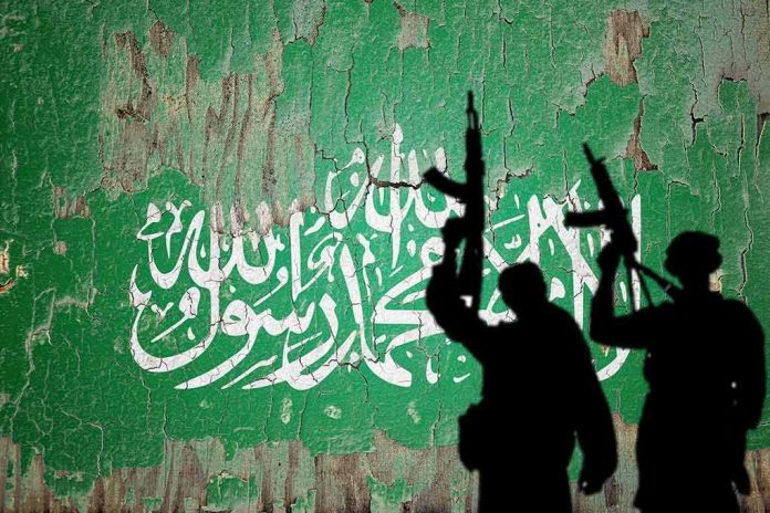 shutterstock_2444744521 silhouettes of armed figures in front of a weathered green wall with white script